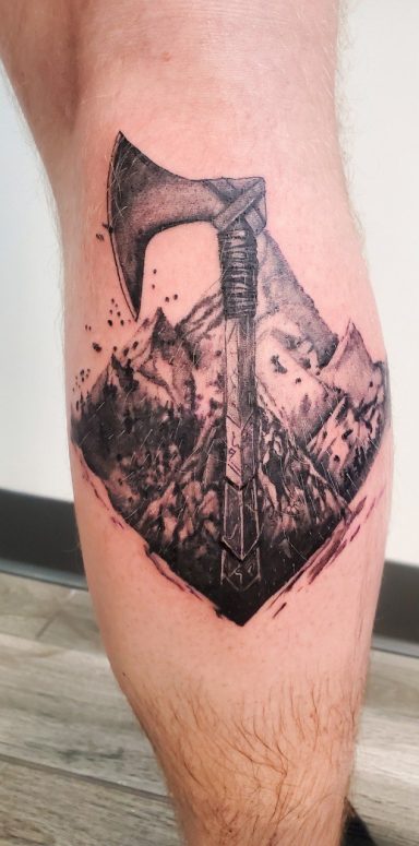 A detailed custom viking style tattoo of an axe set against a rocky mountain background.