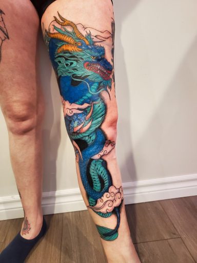 A vibrant custom dragon tattoo wraps around a person's leg, featuring intricate details and colors.